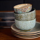 Drie HK Living 70s Ceramics Noodle Kom Verdigris D13-kommen, met blauw-groen gespikkelde patronen, staan gestapeld op bijpassende borden en geven een houten tafel een retro-uitstraling.
