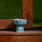 De HK Living 70s Ceramics Tapas Kom op Voet Patina S, met een blauw en bruin gevlekte afwerking, staat op een houten oppervlak bij een houten raam met een groene muur op de achtergrond.
