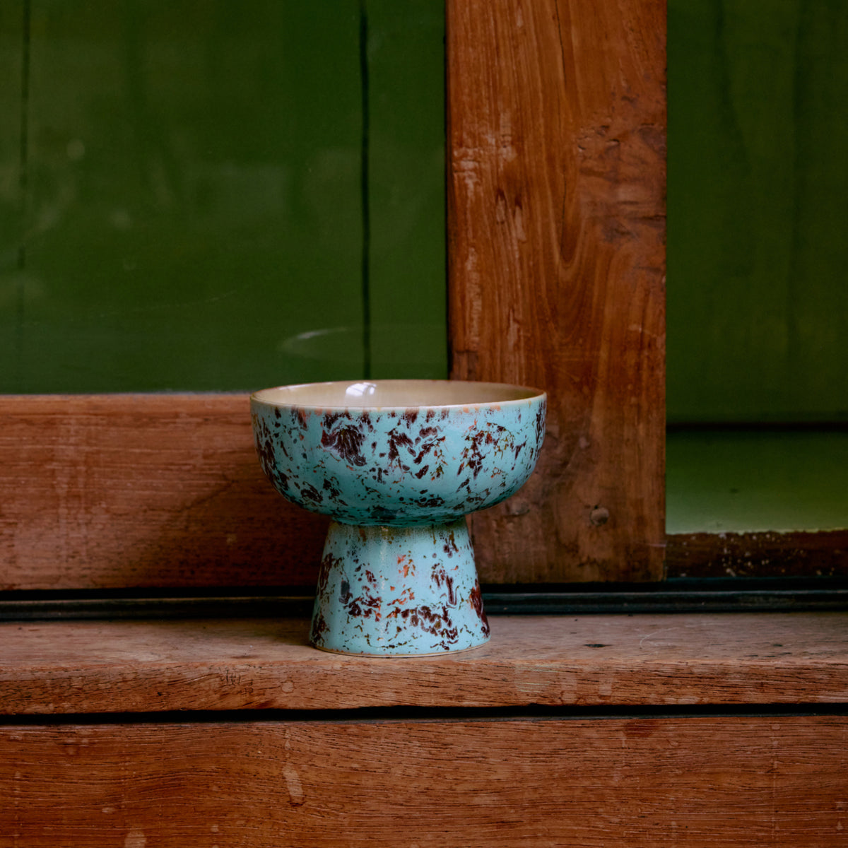 De HK Living 70s Ceramics Tapas Kom op Voet Patina S, met een blauw en bruin gevlekte afwerking, staat op een houten oppervlak bij een houten raam met een groene muur op de achtergrond.