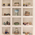 Kleurrijke keramische kommen, kopjes, borden en de HK Living 70s Ceramics Vaasjes Pursuit (5 st.) van HK Living zijn gerangschikt op een witte ingebouwde plank met drie rijen-ideaal voor retro interieur decoratie. Elke nis benadrukt unieke stukken aardewerk.