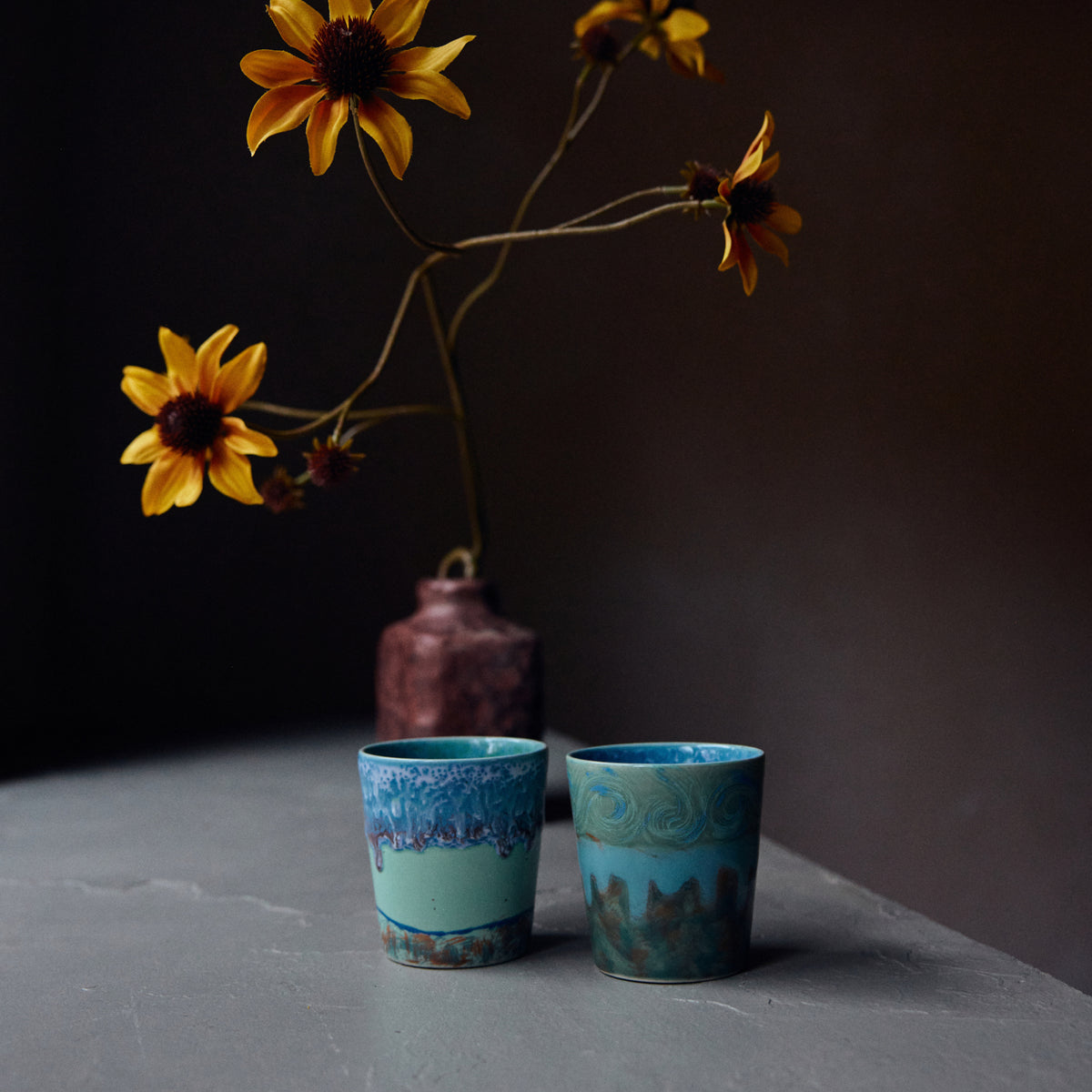 Een keramieken vaas met gele bloemen staat op een grijs oppervlak naast twee HK Living 70s Ceramics Van Gogh Koffiekop Self Portrait mokken. De donkere achtergrond benadrukt dit stijlvolle stilleven, dat vintage charme combineert met moderne flair.