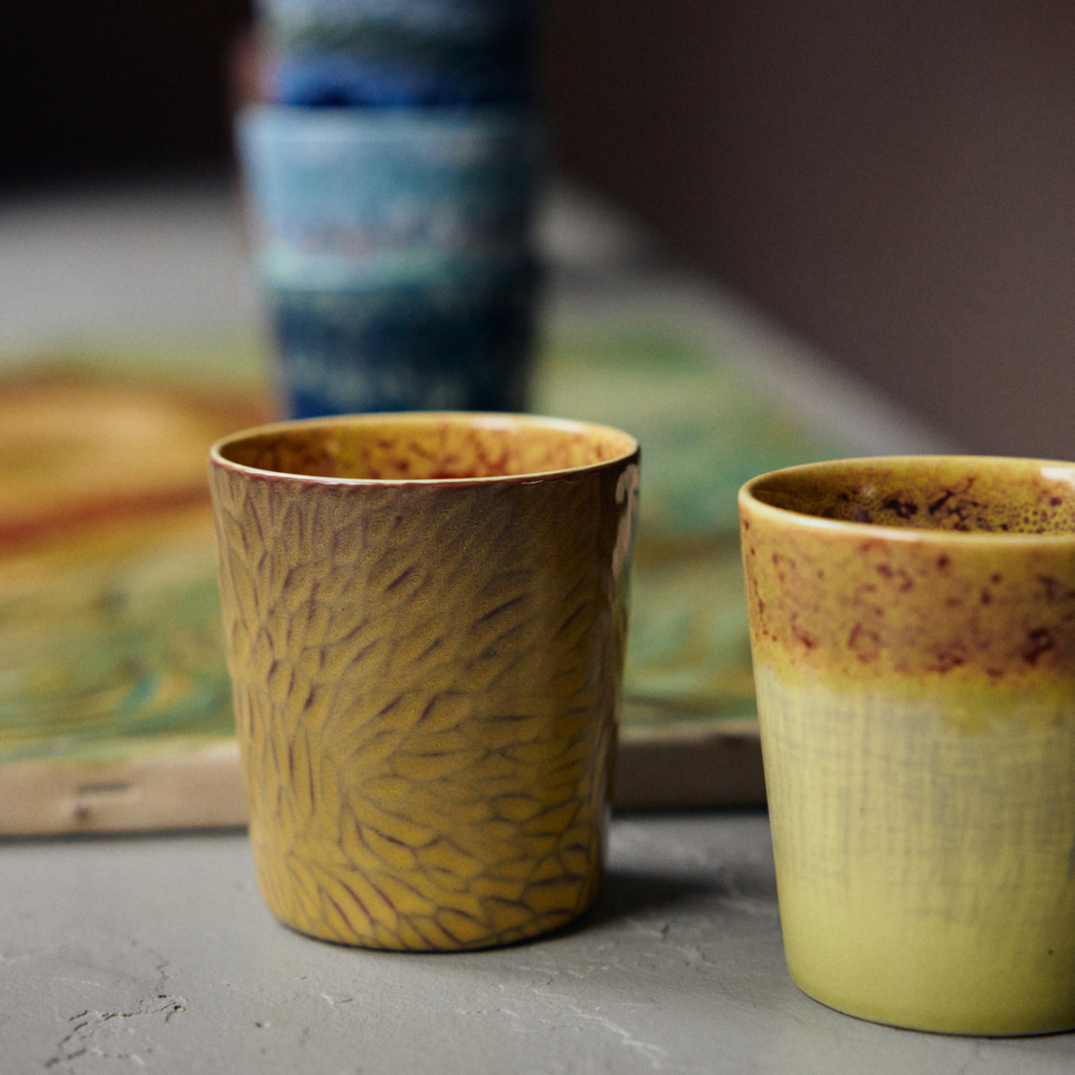Twee HK Living 70s Ceramics Van Gogh Koffiekop Zonnebloemen kopjes met gele en bruine motieven rusten op een grijs oppervlak; de linker heeft een bladmotief met structuur, de rechter een reactief glazuurverloop. Vage blauwe kopjes staan op de achtergrond.