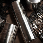 De HK Living Kerstspecial Geurkaarsen Frost & Firewood 3 st. in zilver metallic potjes met zwarte tekst worden getoond naast zwarte veren en een pailletten accessoire-een stijlvol kerstcadeau of om je favoriete geurkaarsen te presenteren.