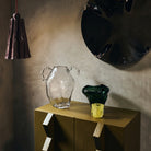 Een stilleven met een gestructureerde vaas van helder glas, een HK Living Coralla Theelichthouder Lime kom op een mosterdkleurige kast, een kastanjebruine hanglamp en een grote zwarte wandsculptuur tegen een grijze muur.