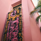 De HK Living Fluffy Tapijt South Beach 360/80 in roze, bruin en geel hangt uit een raam van een roze gebouw, met rechts een palmboom en een lichte lucht, wat de speelse stijl van HK Living weerspiegelt.