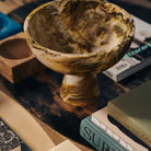 De HK Living gemarmerde Kom op Voet Oker D23, met een bruin en geel gemarmerd ontwerp, staat op een houten tafel versierd met boeken en andere voorwerpen voor een artistieke uitstalling.
