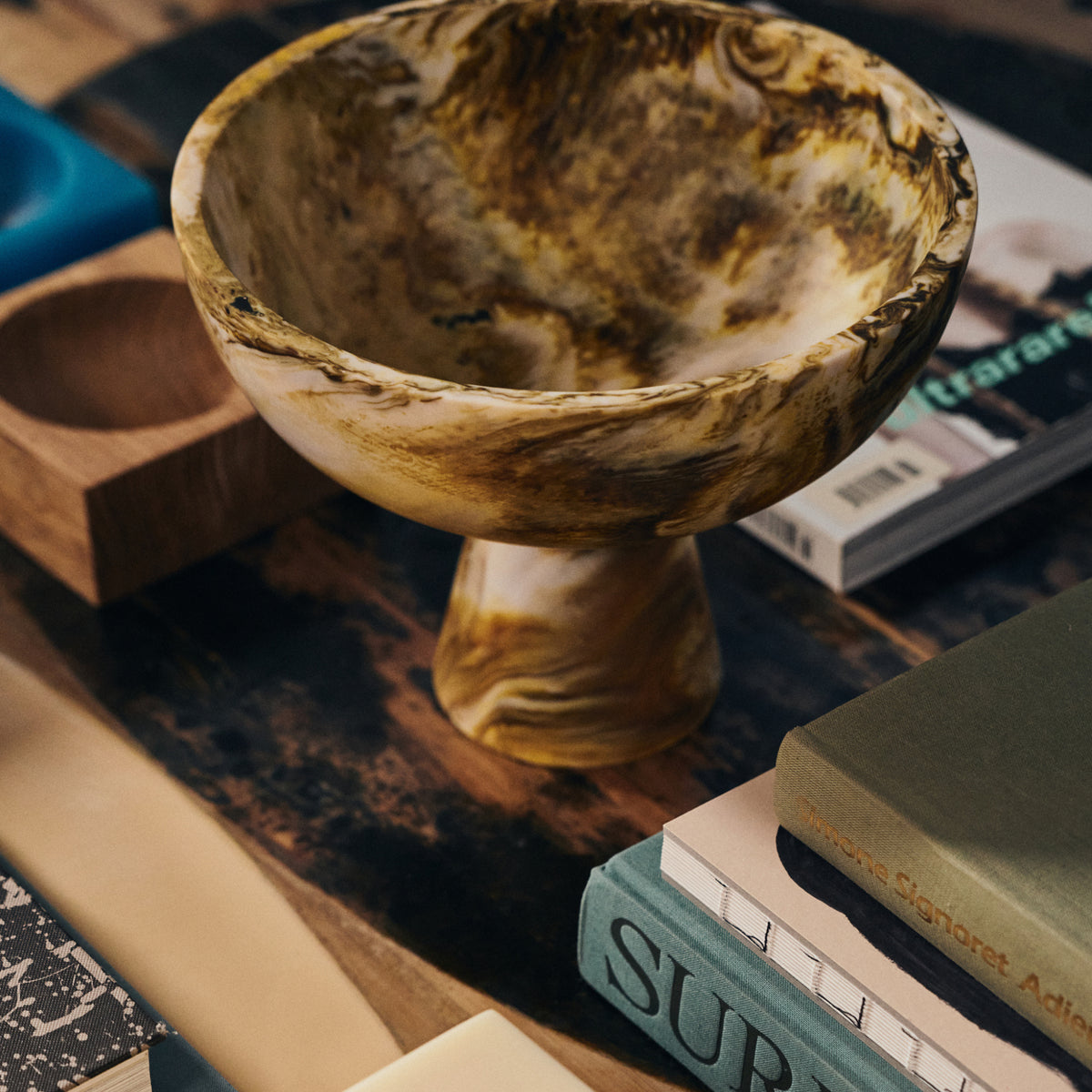 De HK Living gemarmerde Kom op Voet Oker D23, met een bruin en geel gemarmerd ontwerp, staat op een houten tafel versierd met boeken en andere voorwerpen voor een artistieke uitstalling.