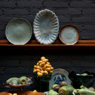 Drie decoratieve borden op een houten plank tegen een donkere bakstenen muur, met de HK Living New Classics Resin Schaal Lichtblauw en diverse groene vruchten op de tafel eronder, die moderne elegantie toevoegen.