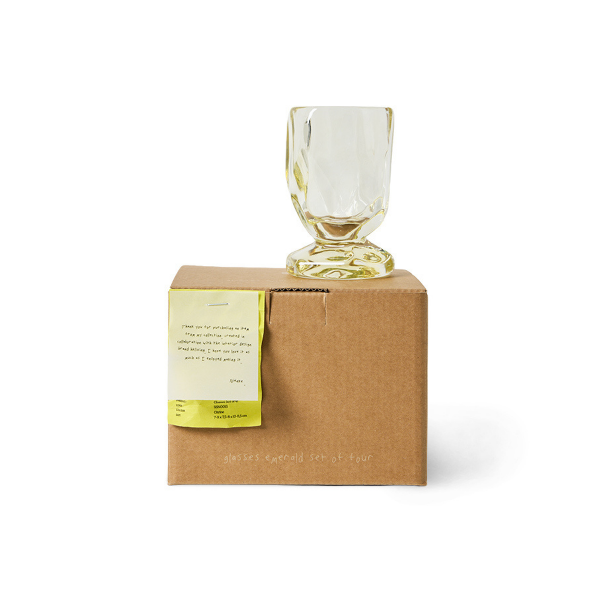 Een enkel helder, gestructureerd glas uit de HK Living Nienke Sikkema Wijnglas Citrine Geel 4-delige set staat ondersteboven op een bruine doos met een geel etiket, waardoor een stijlvol, minimalistisch tafereel ontstaat tegen een witte achtergrond.