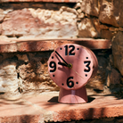 De HK Living Retro Klok Roze, een roze keramische klok met een golvend ontwerp, staat op een zonovergoten bakstenen trede voor een stenen muur. De HK Living-klok geeft 10:10 aan.