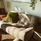 Een beige getufte loveseat met een HK Living Rugged Kussen Olijfgroen 50/50 kussen staat in een zonovergoten hoek, naast houten bijzettafeltjes en een hangende bladplant.