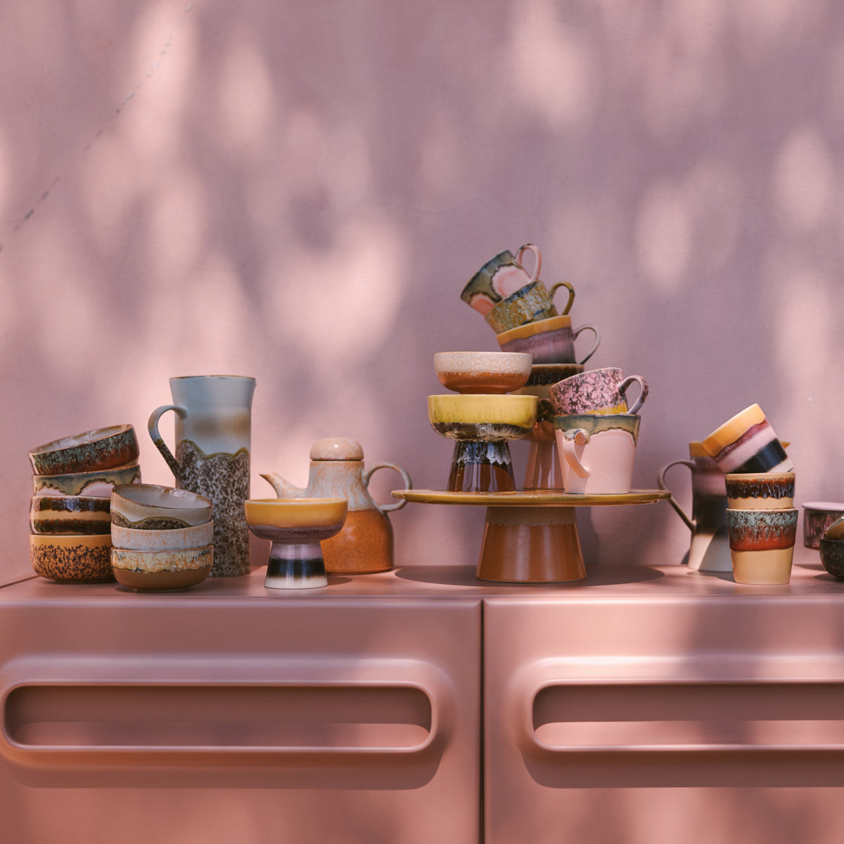 Een stapel HK Living 70s Ceramics Koffiekop Cove mokken geeft een kleurrijk retro tintje aan een roze dressoir tegen een zonovergoten roze muur.