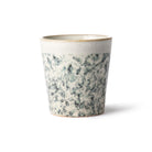 De HK Living 70s Ceramics Koffiekop Hail heeft een licht uitlopende rand en een retro gespikkeld wit en blauwgrijs glazuur, dat de vintage stijl weerspiegelt, afgebeeld tegen een effen witte achtergrond.