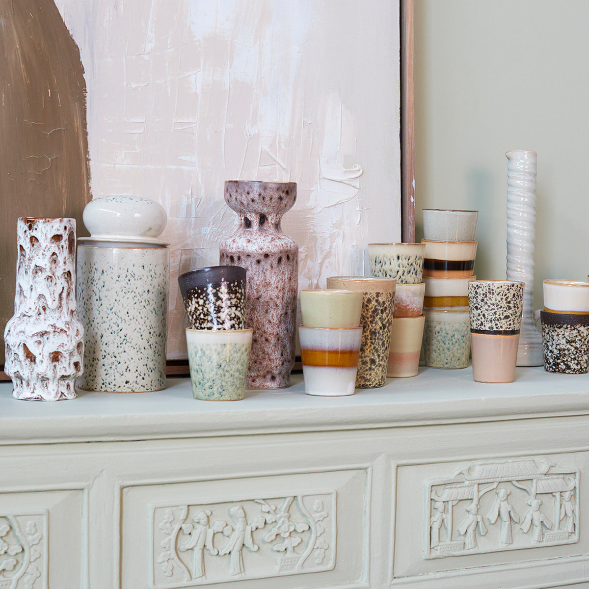 De HK Living 70s Ceramics Koffiekop Hail, met zijn retro gespikkelde en gestreepte ontwerp, staat op een witte schoorsteenmantel voor abstracte kunstwerken.