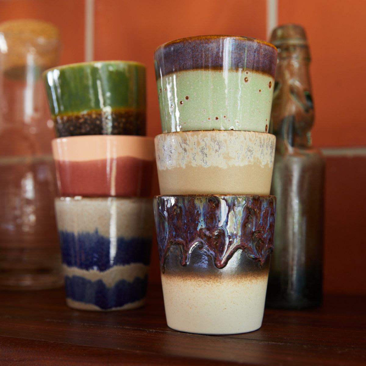 Zes HK Living 70s Ceramics Koffiekop Cosmos kopjes, elk met unieke glazuren en aardse retro tinten, staan op een plank bij een oranje betegelde muur - perfect om vintage flair aan je keuken toe te voegen.