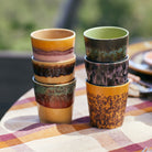 Zes HK Living 70s Ceramics Koffiekop Cove-kopjes, elk met een uniek glazuurpatroon, zijn paarsgewijs gestapeld op een geruit tafelkleed buiten en brengen retrostijl in je omgeving.