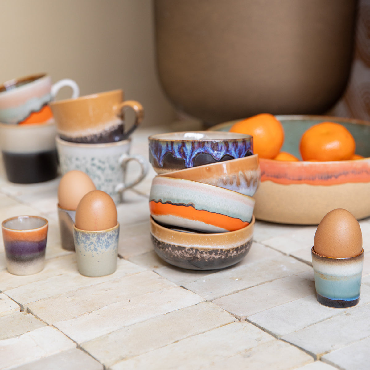 Een set HK Living 70s Ceramics Tapasschaaltje Crystal 4 st. rust op een betegeld oppervlak met kopjes, kommen en bruine eieren; op de achtergrond benadrukt een schaal met sinaasappels de kleurrijke, aardse glazuren.