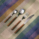 Het HK Living 70s Bestek Tafellepel Hout van HK Living heeft een theelepel, eetlepel, vork en mes met houten handvatten, naast elkaar uitgestald op een gestreept, veelkleurig tafelkleed.