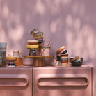 Een stapel HK Living 70s Ceramics Koffiekop Cliffs kopjes staat op een roze kast tegen een roze muur, met zacht gedimd zonlicht dat retro stijl schaduwen toevoegt aan het kleurrijke tafereel.