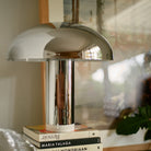 De HK Living Lounge Tafellamp Chroom, een moderne koepelvormige lamp van HK Living, ligt op een stapel boeken voor ingelijste kunstwerken en een groene bladplant.