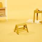 Een Hasegawa Lucano Ladder 1 Step Geel van Hasegawa, geïnspireerd op een minimalistisch ontworpen opstapje, staat op een gele vloer in een kamer met een gele tafel, een mok, een pot en een plank met boeken; ook de achtergrond is geel.