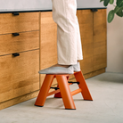 Een persoon in een witte broek staat op een Hasegawa Lucano Ladder 1 Step Oranje van Hasegawa in een keuken met houten kasten en een plant ernaast.