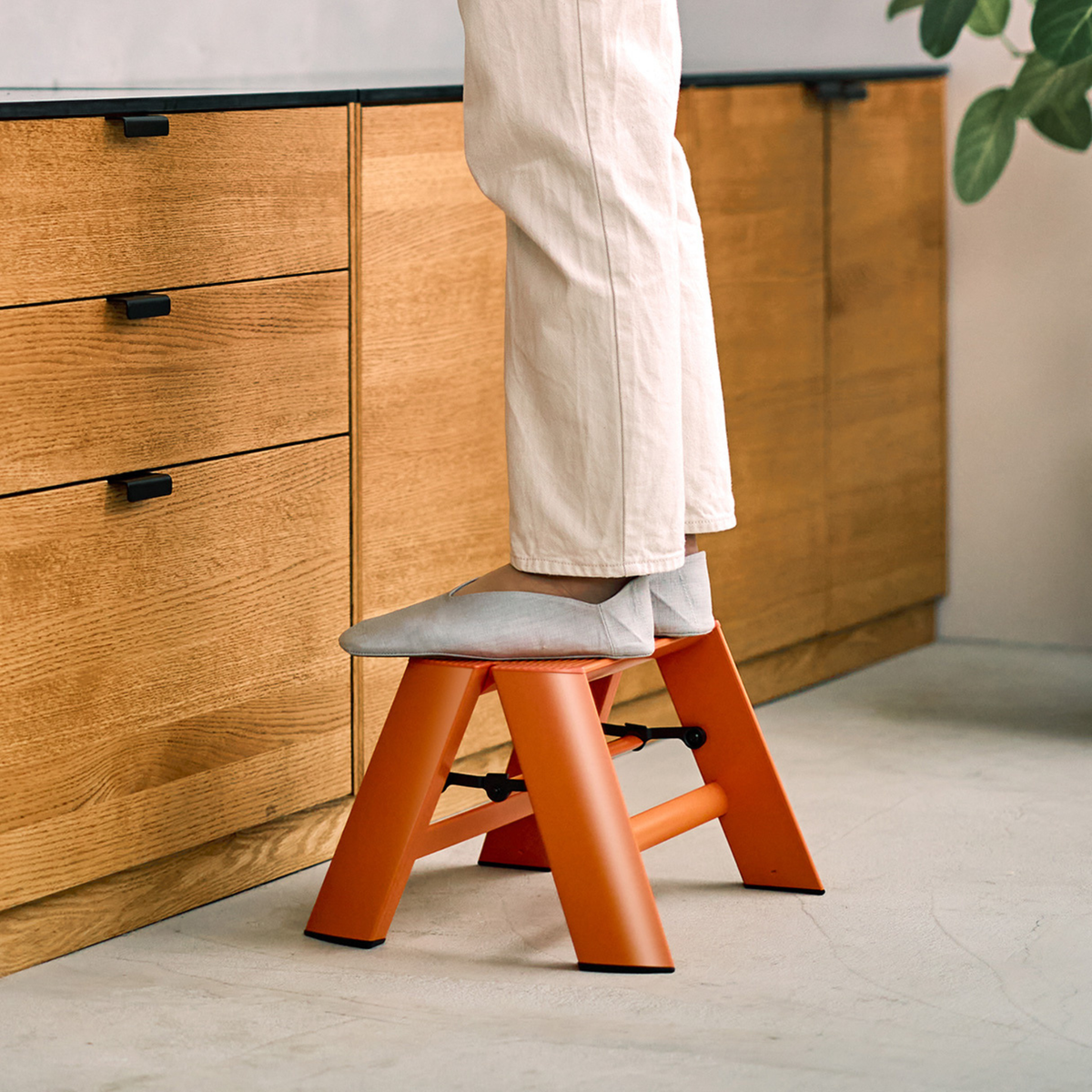 Een persoon in een witte broek staat op een Hasegawa Lucano Ladder 1 Step Oranje van Hasegawa in een keuken met houten kasten en een plant ernaast.