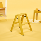 Een Hasegawa Lucano Ladder 2 Step Geel van Hasegawa staat in een kamer waar de muren, de vloer en het meubilair – waaronder een tafeltje met een kopje en boeken op een plank – allemaal geel zijn, waardoor een monochroom geel thema ontstaat dat is geïnspireerd op Japans design.
