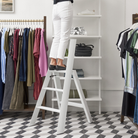 Gekleed in een witte broek beklimt iemand de Hasegawa Lucano Ladder 3 Step Wit Breed van Hasegawa om bij een hoge plank te komen in een kledingwinkel met kledingrekken, een vloer van geometrische tegels en subtiele Japanse designinvloeden in de hele ruimte.