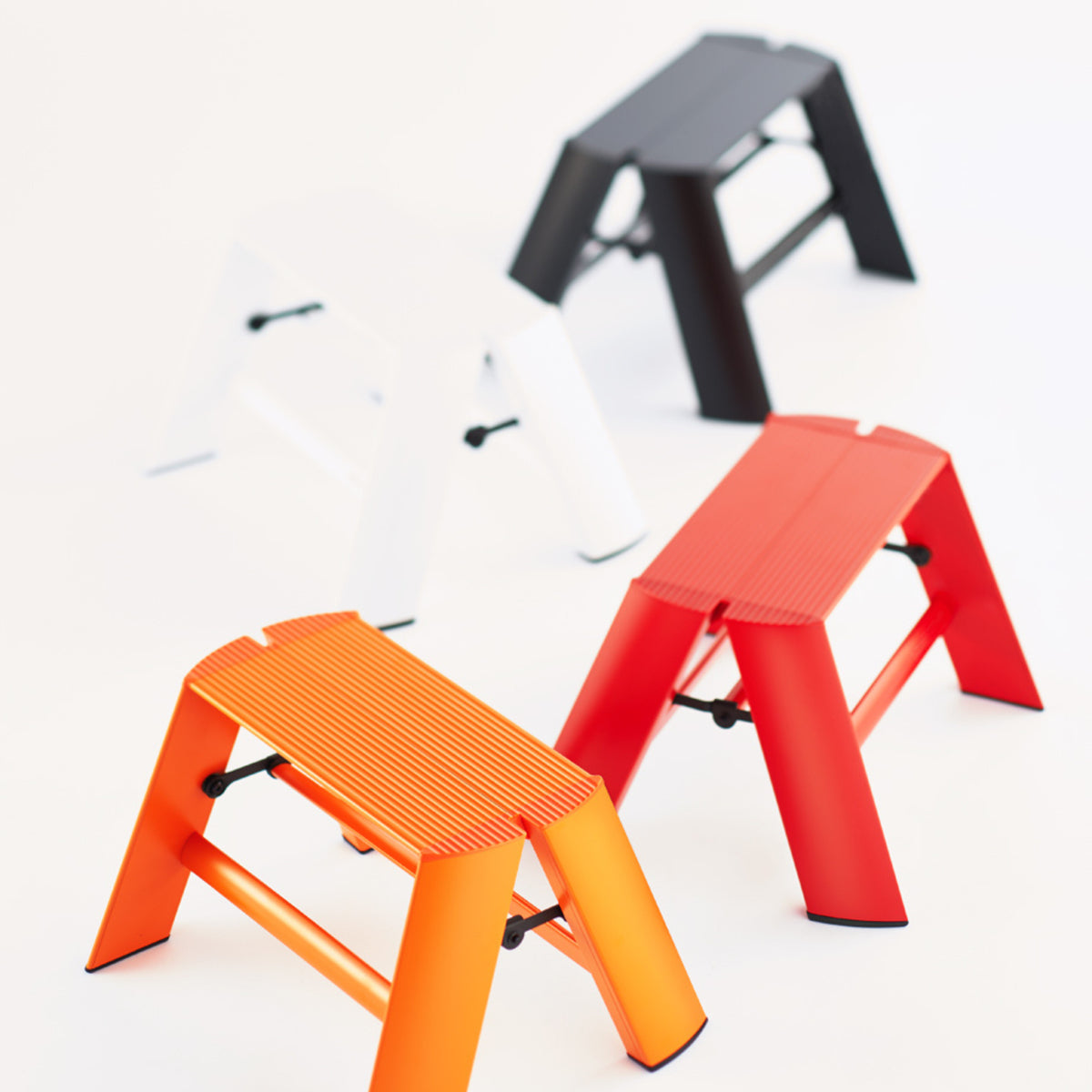 Vier compacte Hasegawa Lucano Ladder 1 Step krukjes in oranje, rood, wit en zwart staan op een witte achtergrond. Elk minimalistisch opstapje van Hasegawa heeft schuine poten en geribbelde zittingen voor stijl en stabiliteit.