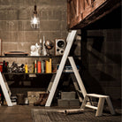 Een gezellige werkkamer in industriële stijl met een houten bureau, boeken, decor, luidspreker, modelraket, laarzen, opbergdozen en een Hasegawa Lucano Ladder 1 Step Wit van Hasegawa ligt op een vloerkleed onder warme hanglampen.