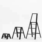 Drie zwarte Hasegawa Lucano Ladder 1 Stap Zwart ladders van Hasegawa staan in een rij op een effen witte achtergrond, waardoor hun compacte Japans geïnspireerde ontwerp en variërende hoogtes van de kortste naar de hoogste benadrukt worden.