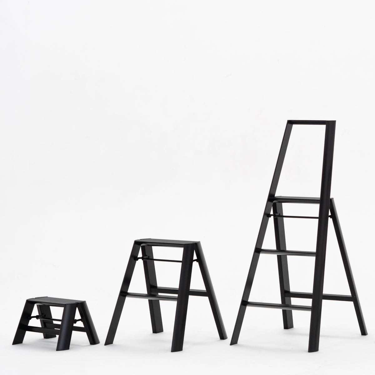 Drie zwarte Hasegawa Lucano Ladder 1 Stap Zwart ladders van Hasegawa staan in een rij op een effen witte achtergrond, waardoor hun compacte Japans geïnspireerde ontwerp en variërende hoogtes van de kortste naar de hoogste benadrukt worden.