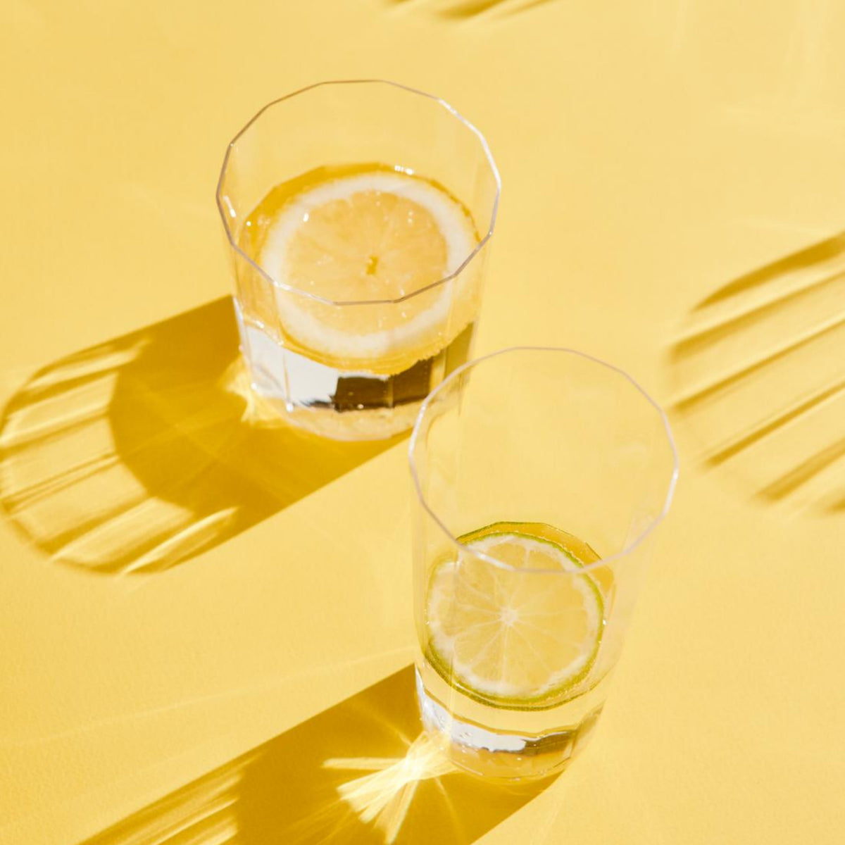 Twee Hay Angle Waterglazen gevuld met schijfjes citroen werpen scherpe schaduwen op een helder geel oppervlak. De sterke verlichting versterkt hun elegante look, creëert levendige reflecties en een fris, zomers gevoel.