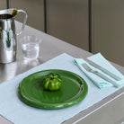 Een enkele groene tomaat rust op een Hay Barro Borden Groen D24 van Hay op een lichtblauwe placemat, met een vork en mes op een bijpassend servet vlakbij, plus een glas water en een zilveren kan op de roestvrijstalen tafel.
