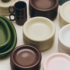 Stapels keramische borden - waaronder Hay Barro Borden Bordeaux D18 van Hay - in bruin, crème, groen, wit en roze staan op een tafel met bruine mokken en ovale groene serveerschalen.