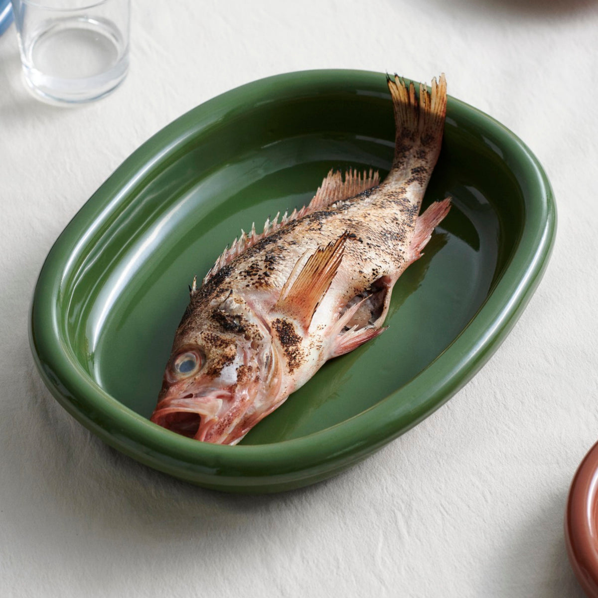 Een hele rauwe vis met open bek ligt in een Hay Barro Ovale Schaal Groen 27/36 van Hay, onderdeel van de terracotta collectie, op een wit tafelkleed met wat water; vlakbij staan een glas en een bord, die de elegante stijl van Rui Pereira oproepen.