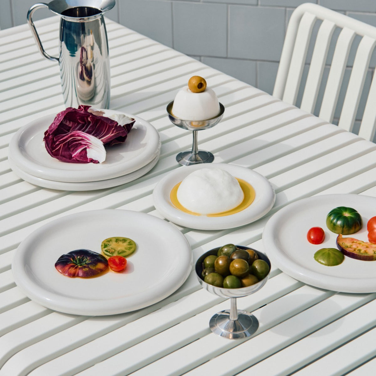 Een Hay Barro Borden Off-White D24 bordenset van Hay bevat borden met kleurrijke tomaten, paarse sla, groene olijven, een dessert met een olijf erop, een metalen kan en een schaal met wit schuim en gele saus tegen betegelde muren.