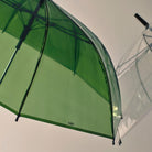 Een close-up van de Hay Canopy Paraplu Groen, met het HAY-logo, zit naast een transparante paraplu, beide vastgelegd tegen een eenvoudige, lichte achtergrond.