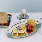 Op een minimalistisch zilveren dienblad liggen partjes geschilde grapefruit, sinaasappel en bloedsinaasappel. Op de achtergrond staat een Hay Essential Steel Eierdop van Hay met daarin een ei, naast een lepel en een bord met gesneden brood, allemaal opgesteld op een wit tafelkleed.