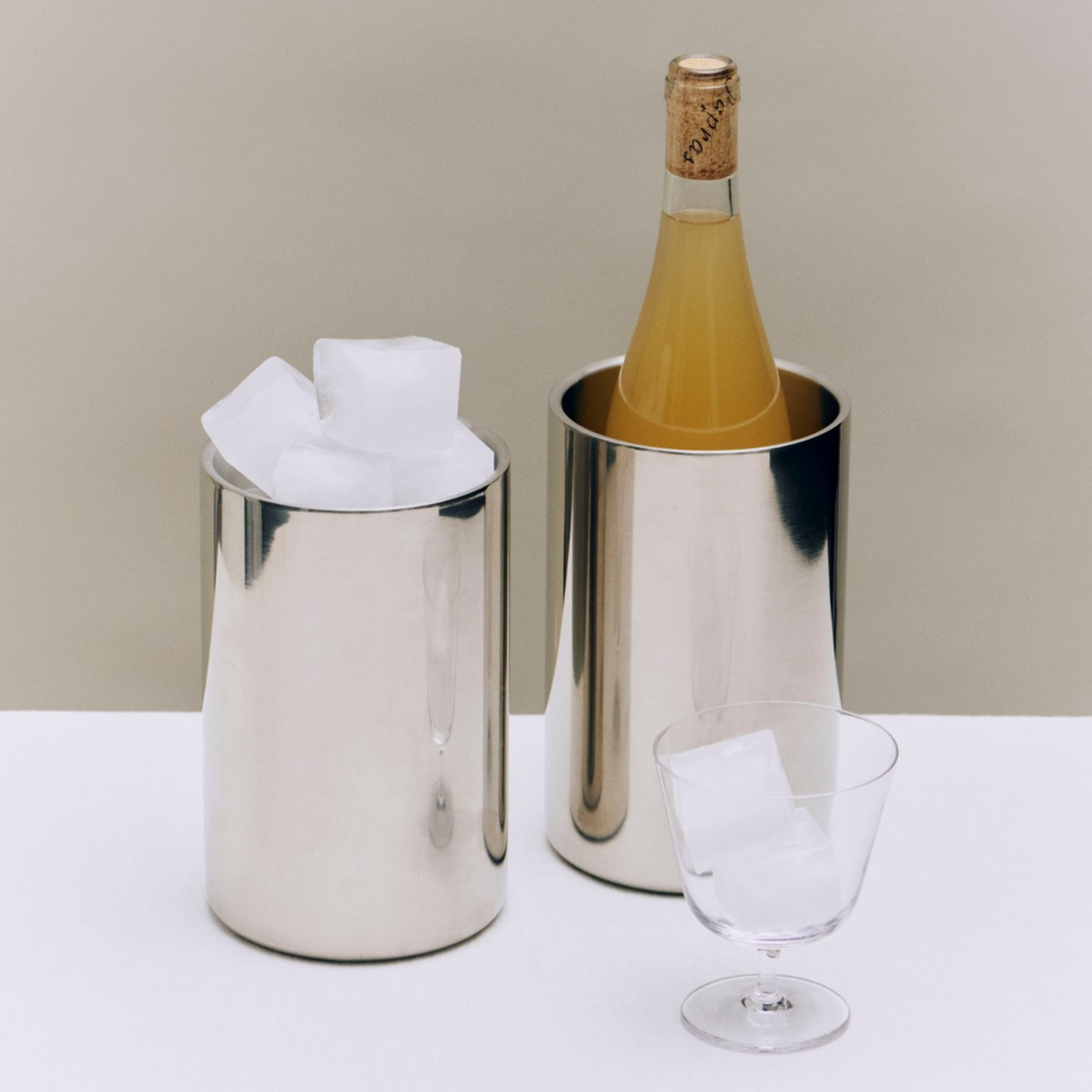 Twee ‘Essential Steel Wijnkoeler’-ijskuipjes van Hay – een strak exemplaar met daarin een fles met kurk en een ander gevuld met ijsblokjes – staan op een wit oppervlak naast een leeg glas met ijsblokjes.