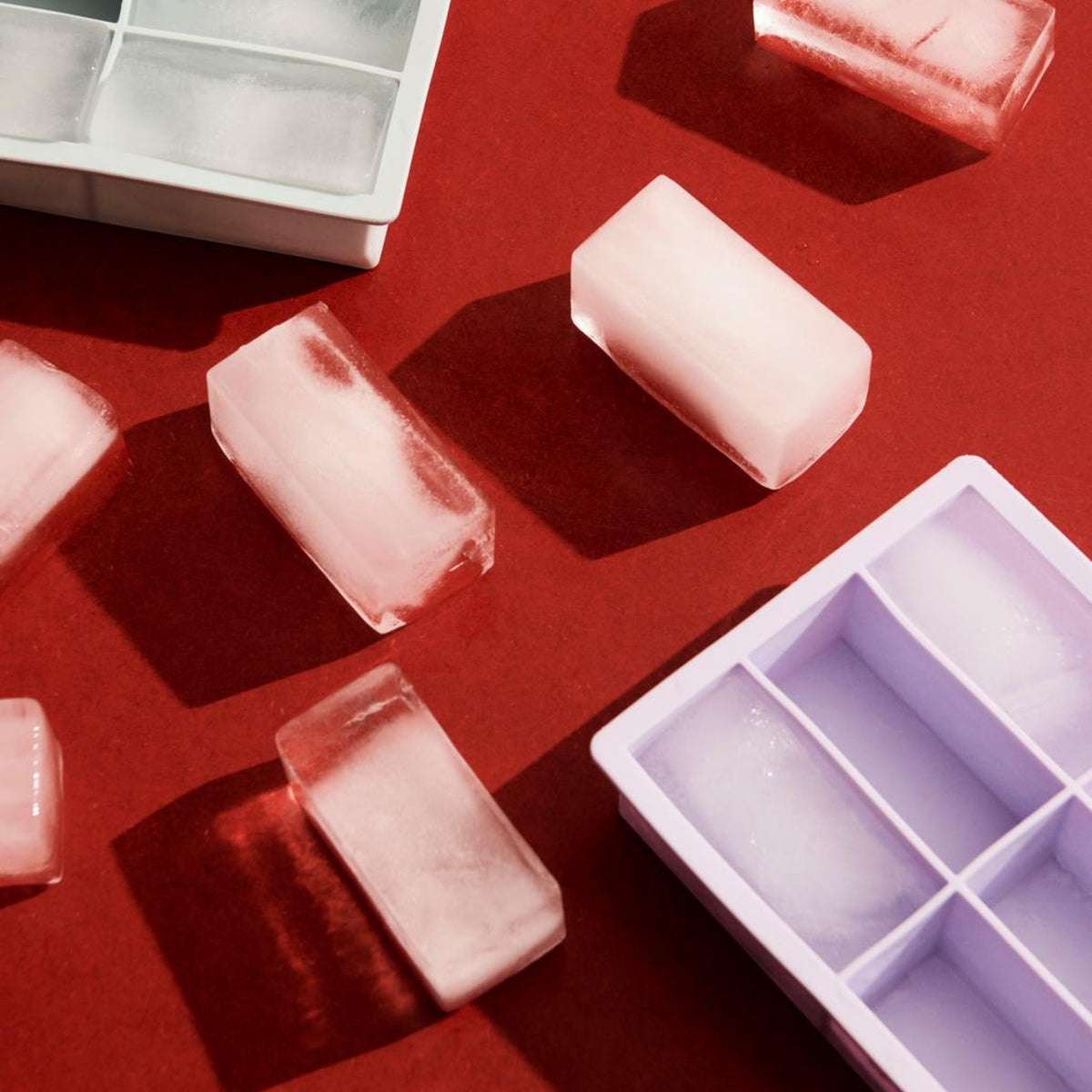 De Hay ijsblokjesvorm Rechthoekig XXL Ijsblauw ijsblokjes worden weergegeven op een rood oppervlak naast witte en paarse siliconen bakjes, met schaduwen die duiden op heldere, directe verlichting.