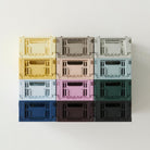 Twaalf kleurrijke opbergkratten, waaronder de Hay Kleur Krat Mini Lichtblauw van Hay, staan in een raster van 3x4 tegen een witte muur. Ze hebben allemaal open handgrepen en een geribbeld ontwerp, gemaakt van gerecycled plastic.