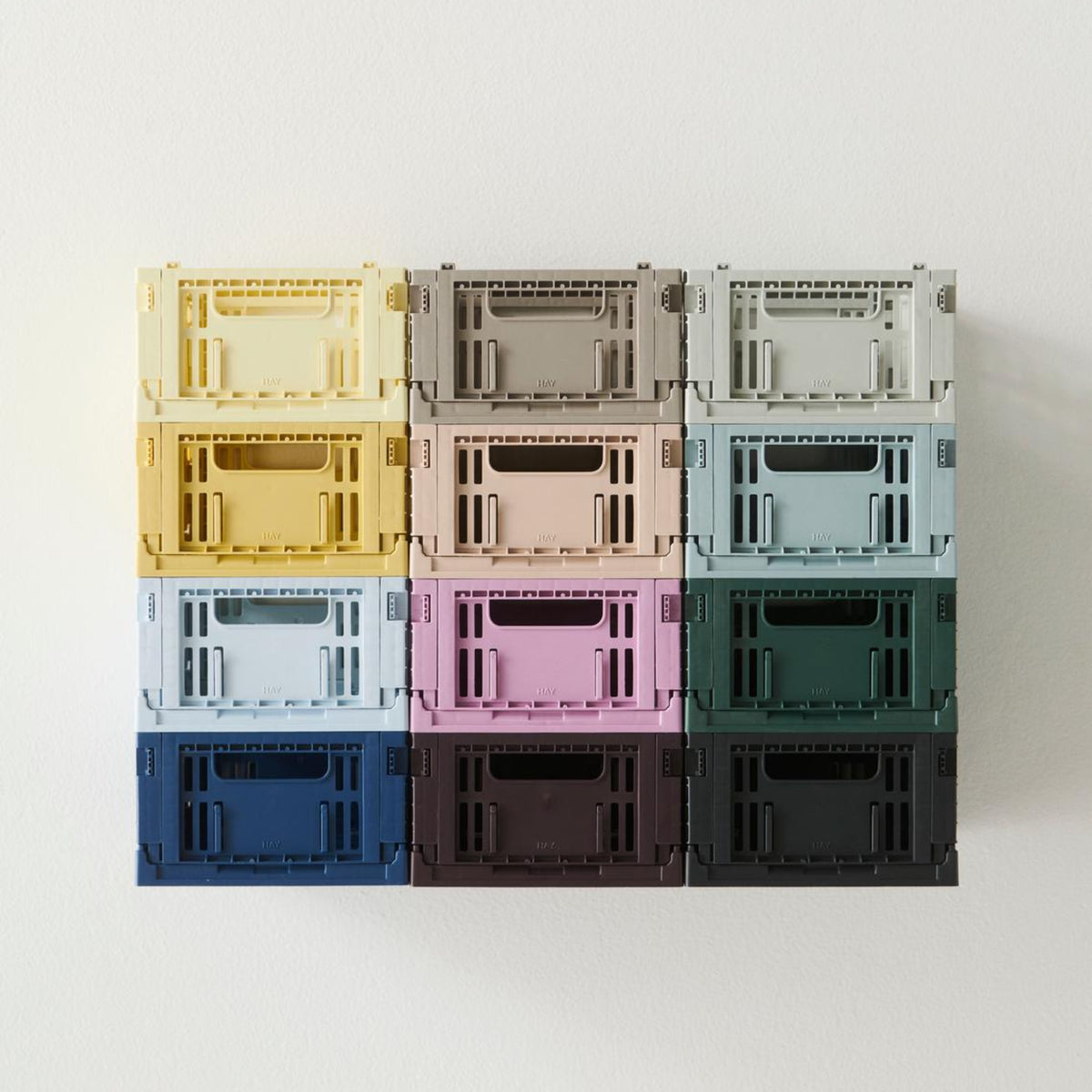 Twaalf kleurrijke opbergkratten, waaronder de Hay Kleur Krat Mini Lichtblauw van Hay, staan in een raster van 3x4 tegen een witte muur. Ze hebben allemaal open handgrepen en een geribbeld ontwerp, gemaakt van gerecycled plastic.