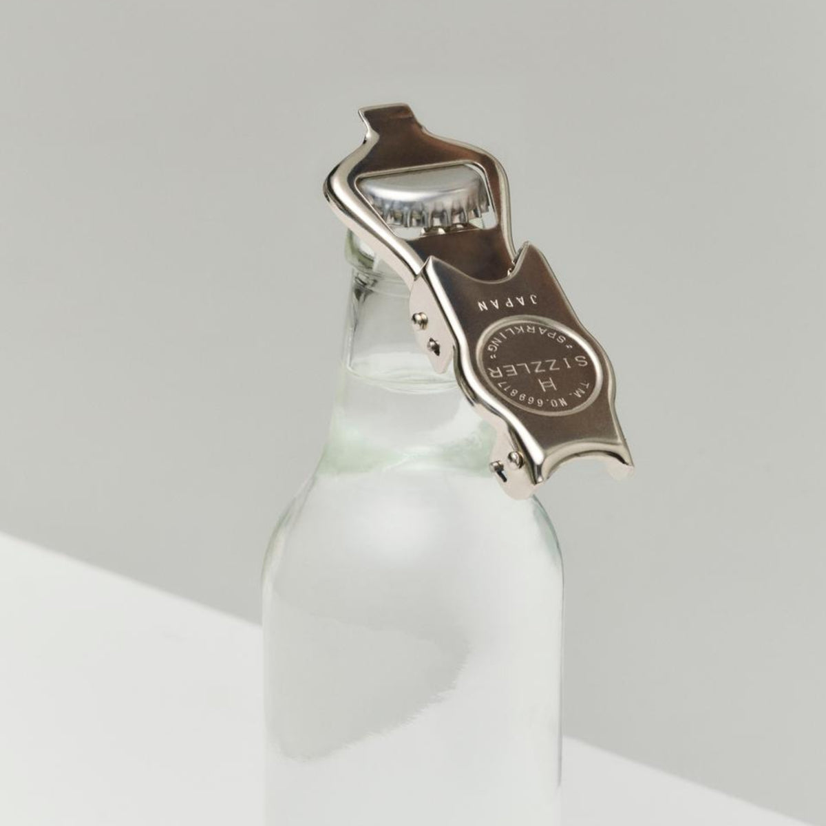 Een Hay Outdoor Market Flesopener van Hay rust bovenop een heldere glazen fles, klaar om te openen. De minimalistische achtergrond benadrukt de opener en de fles - ideaal voor buitenactiviteiten.