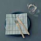 Een blauw bord met Hay Pattern Papieren Servetten Lichtblauw Blauw Grid 25/25 van Hay, met daarop vork en mes. Vlakbij staat een helder glas water op een grijsblauw oppervlak.