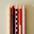 De Hay Patroon Kaarsen Rood en Bordeaux set bestaat uit vier elegante kaarsen met geometrische patronen in rood en bordeaux, naast elkaar gerangschikt op een lichtbeige achtergrond voor een stijlvolle touch.