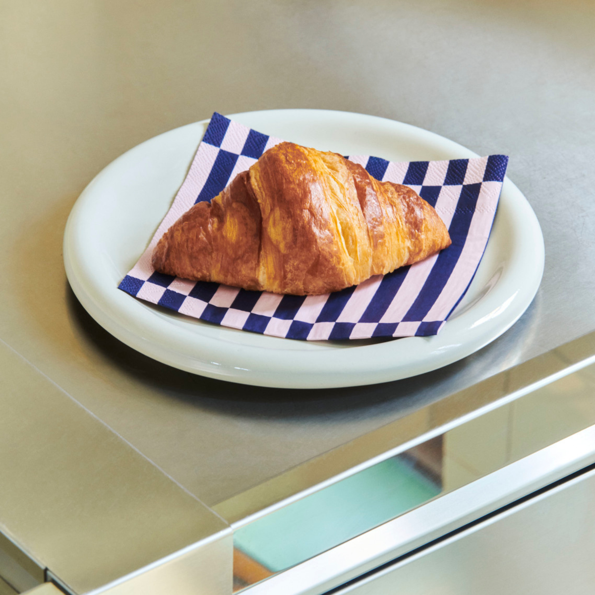 Een gouden croissant rust op een Hay Pattern Papieren Servetten Lichtroze Donkerblauw 33/33 servet van Hay, bovenop een wit bord op een glanzend metalen aanrecht.