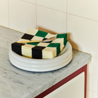 Een stapel Hay Pattern Papieren Servetten Off-White Groen Zwart 33/33 van het merk Hay ligt op drie witte borden op een marmeren aanrecht naast een betegelde muur.