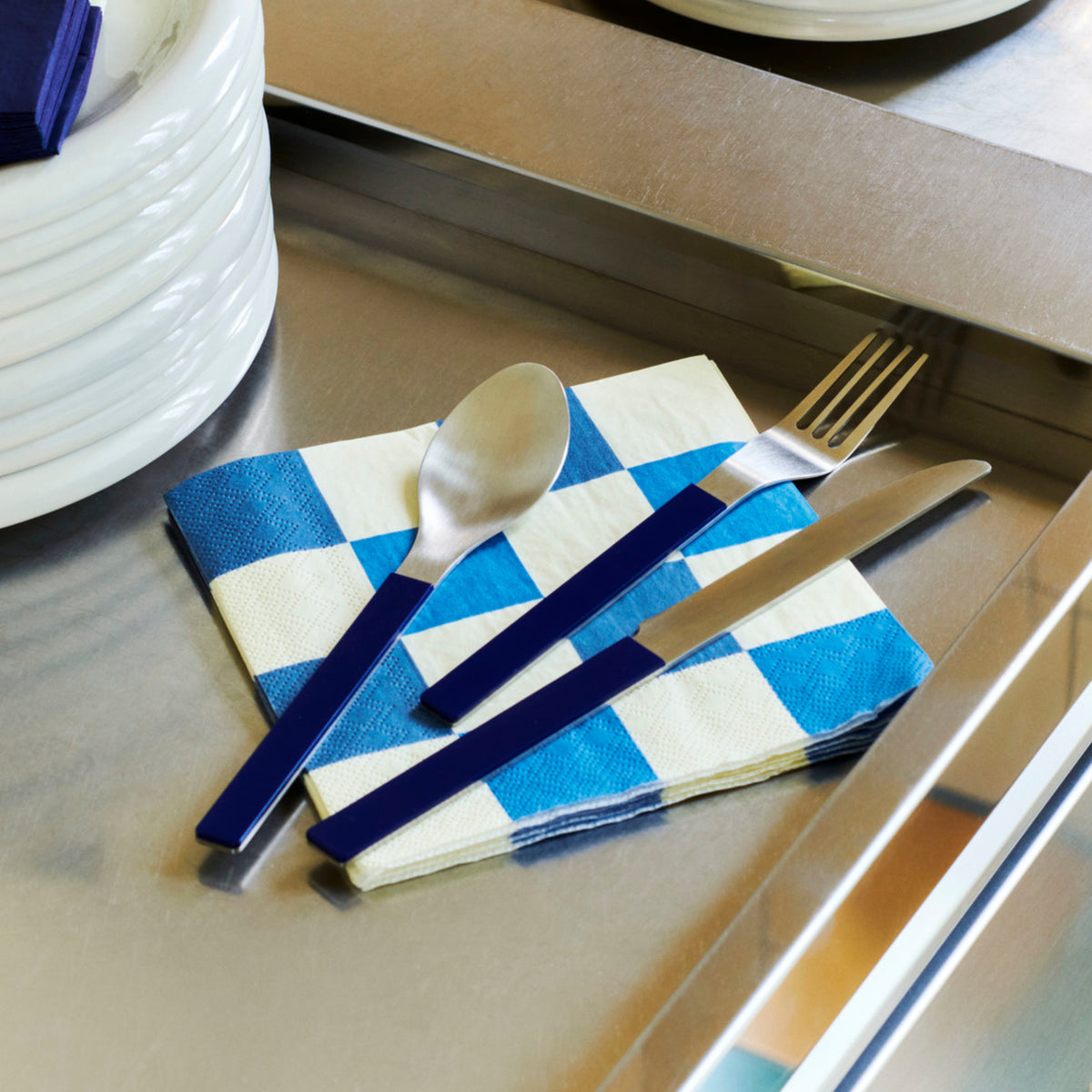 Een lepel, vork en mes met blauwe handvatten rusten op Hay Pattern Papieren Servetten Off-white Blauw Check 33/33 van Hay op een metalen oppervlak naast een stapel witte borden.